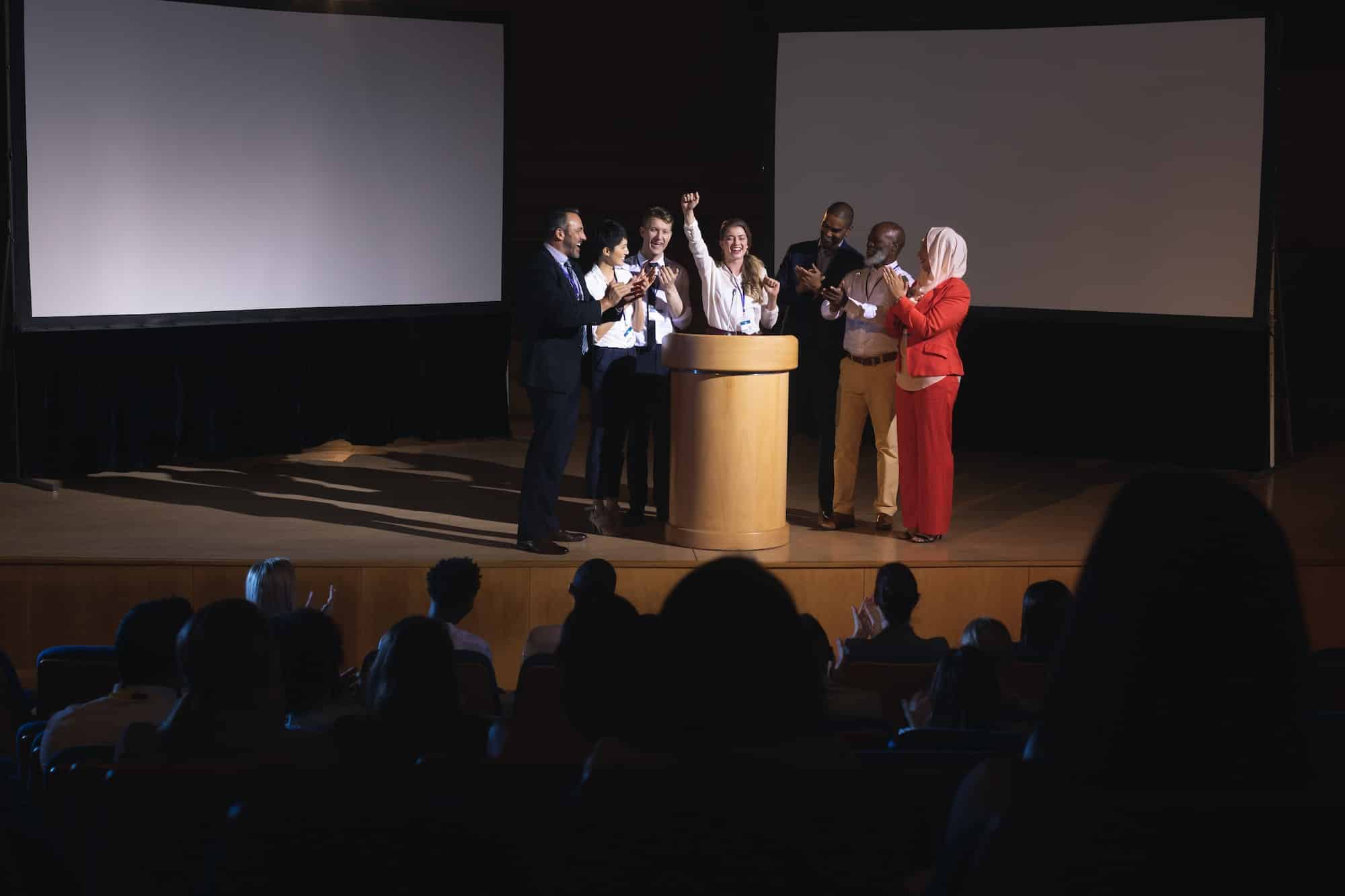 businesswoman-standing-at-the-stage-of-the-auditorium-with-colleagues-in-front-of-audience.jpg